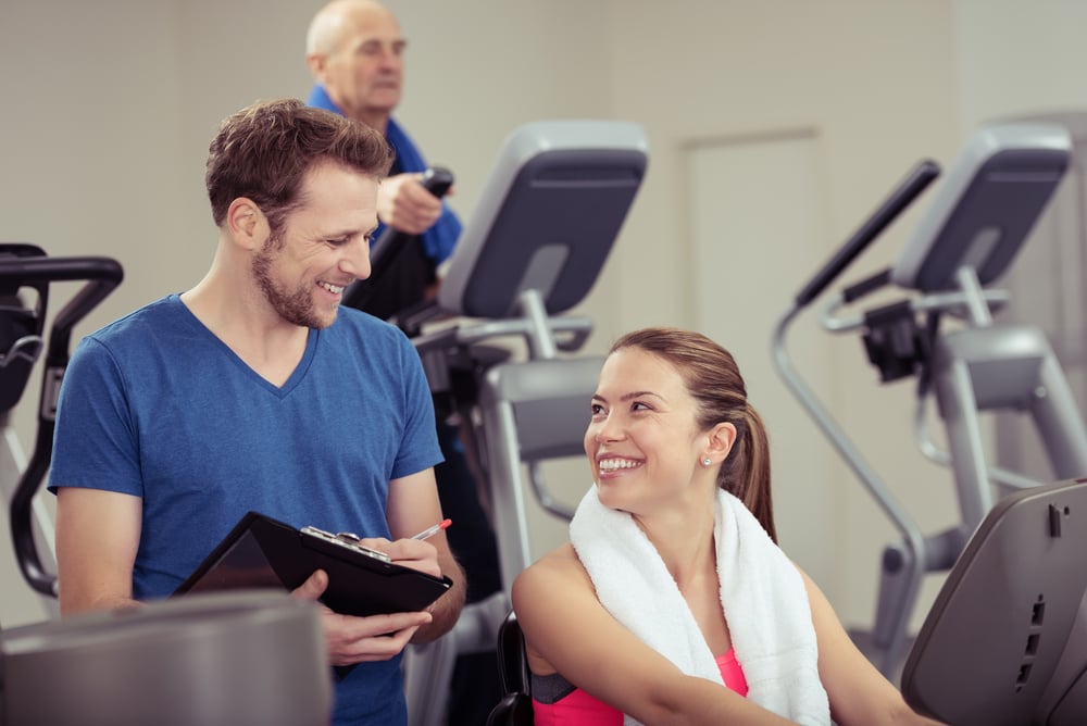 Attractive young woman with a trainer at the gym smiling up at him as they discuss her progress in a health and fitness concept Attractive young woman with a trainer at the gym smiling up at him as they discuss her progress in a health and fitness concept